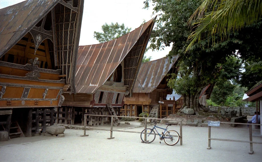 A row of Batak houses on posts with tall saddle-shaped roofs and carved front panels in a village yard