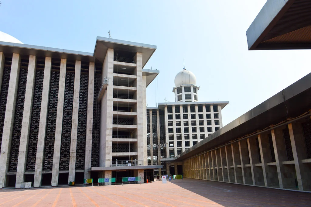 Istiqlal Mosque's courtyard with tall stone-clad facades, patterned screens, a long colonnade and the minaret topped by a white dome