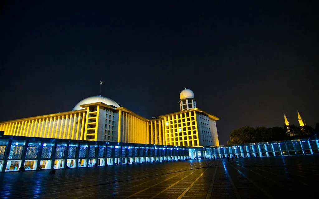 Istiqlal Mosque lit at night with its main dome and minaret, and the twin spires of Jakarta Cathedral visible beyond the trees on the right