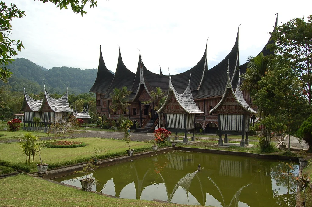 A large rumah gadang with several upswept gonjong roof peaks, with smaller raised rangkiang rice barns and a pond in front